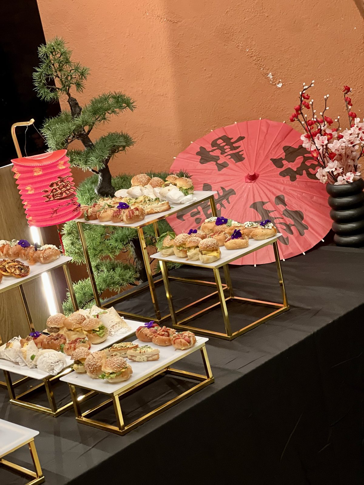Buffet salé installé pour une réception à thème asiatique, présentoirs dorés garnis de mini burgers et sandwichs